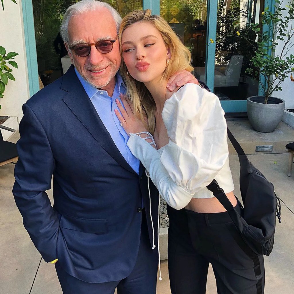 Nicola Peltz Beckham, in a cropped white shirt and black pants, hugs Nelson Peltz in a blue suit and blue shirt.