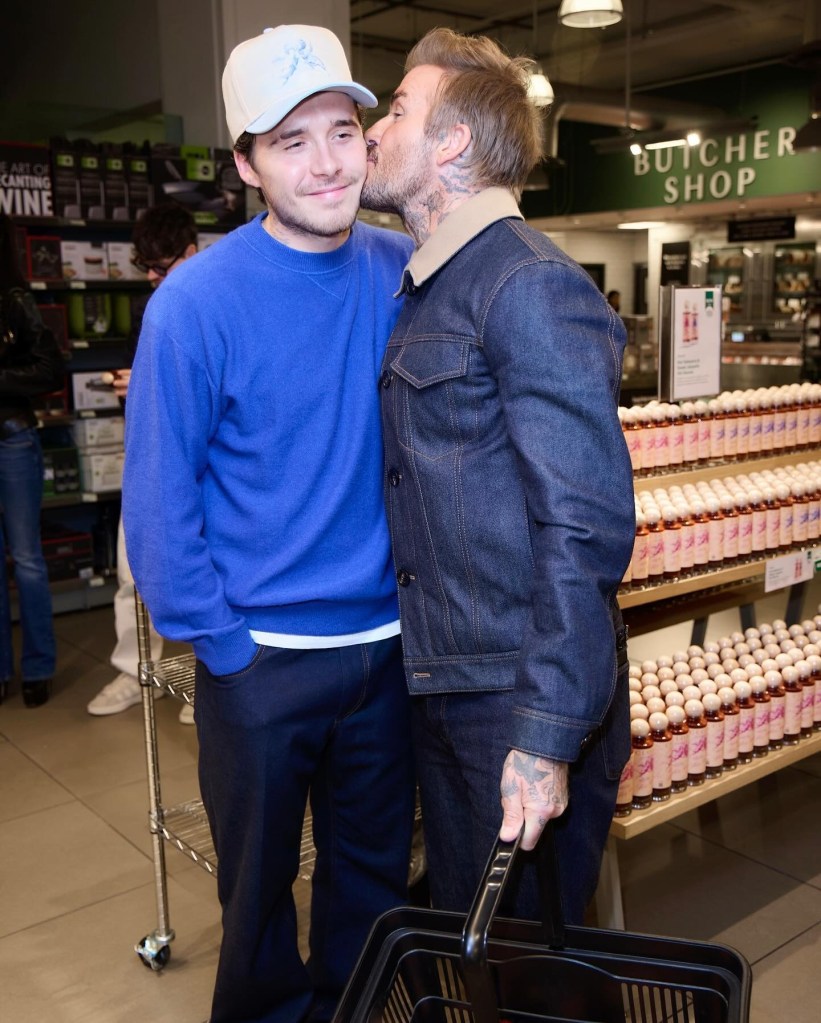 David Beckham in a denim jacket and black pants, Brooklyn Beckham in a blue sweater, white baseball cap and dark pants.