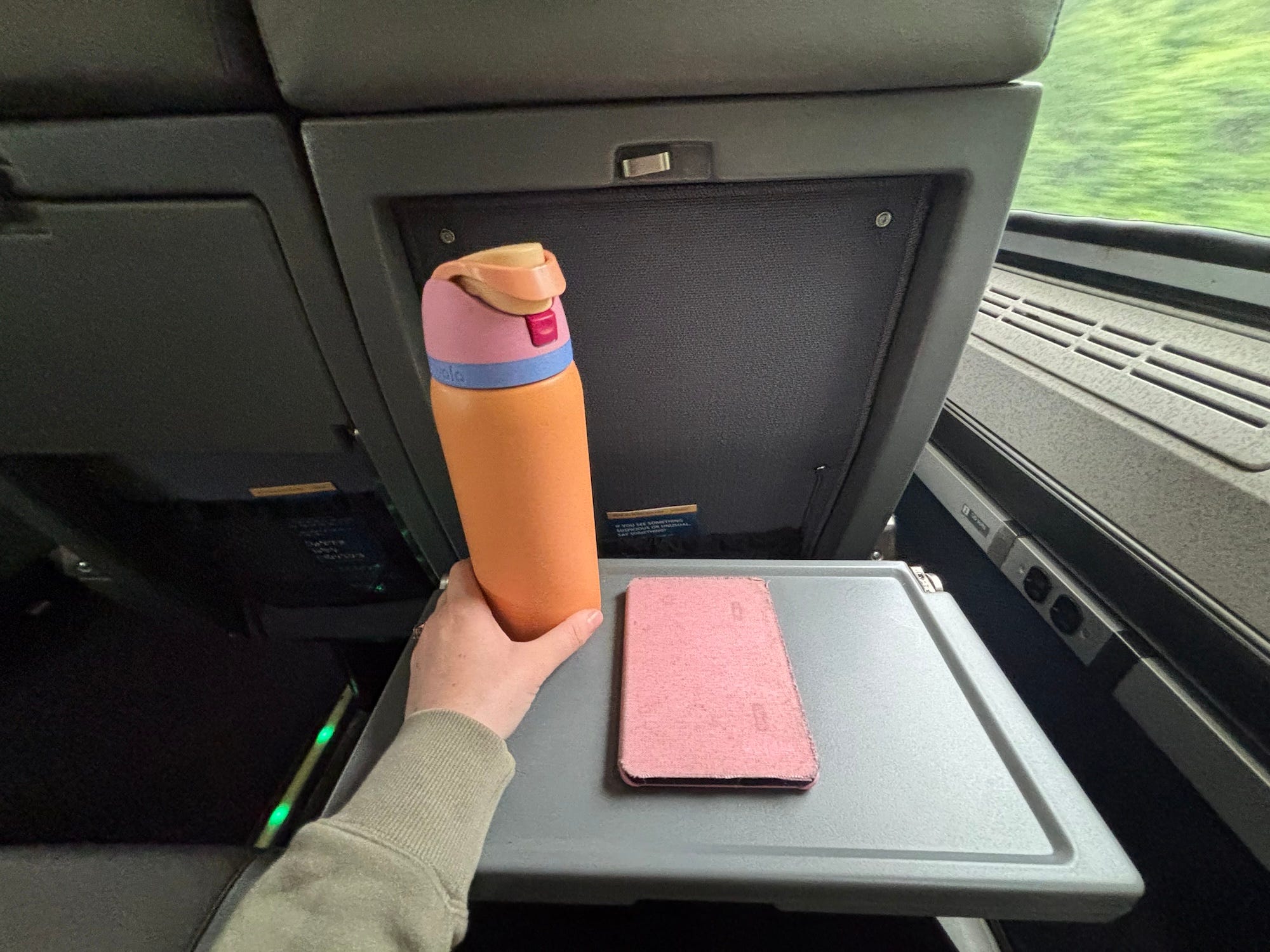 An open tray on the back of a train seat. An orange water bottle and pink kindle sit on the table, and a woman's hand reaches for the bottle.