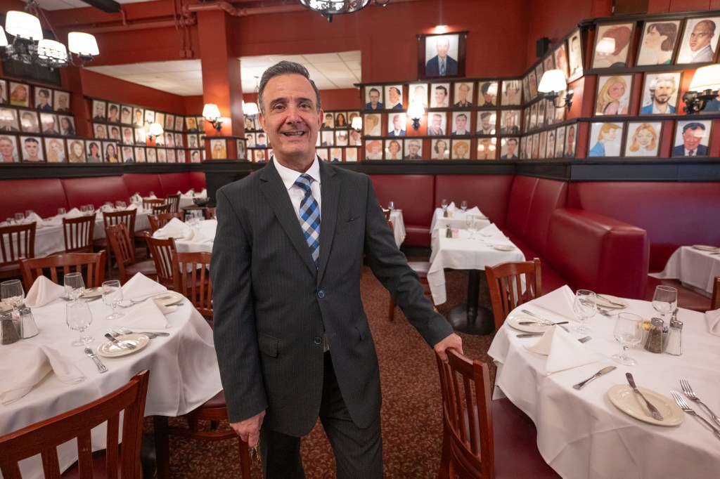 Maitre d' Johnny Felidi at Sardis standing in a restaurant with caricature-filled walls.