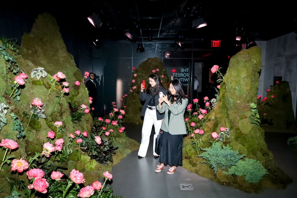 Two women taking photos in a room decorated with moss and pink flowers.