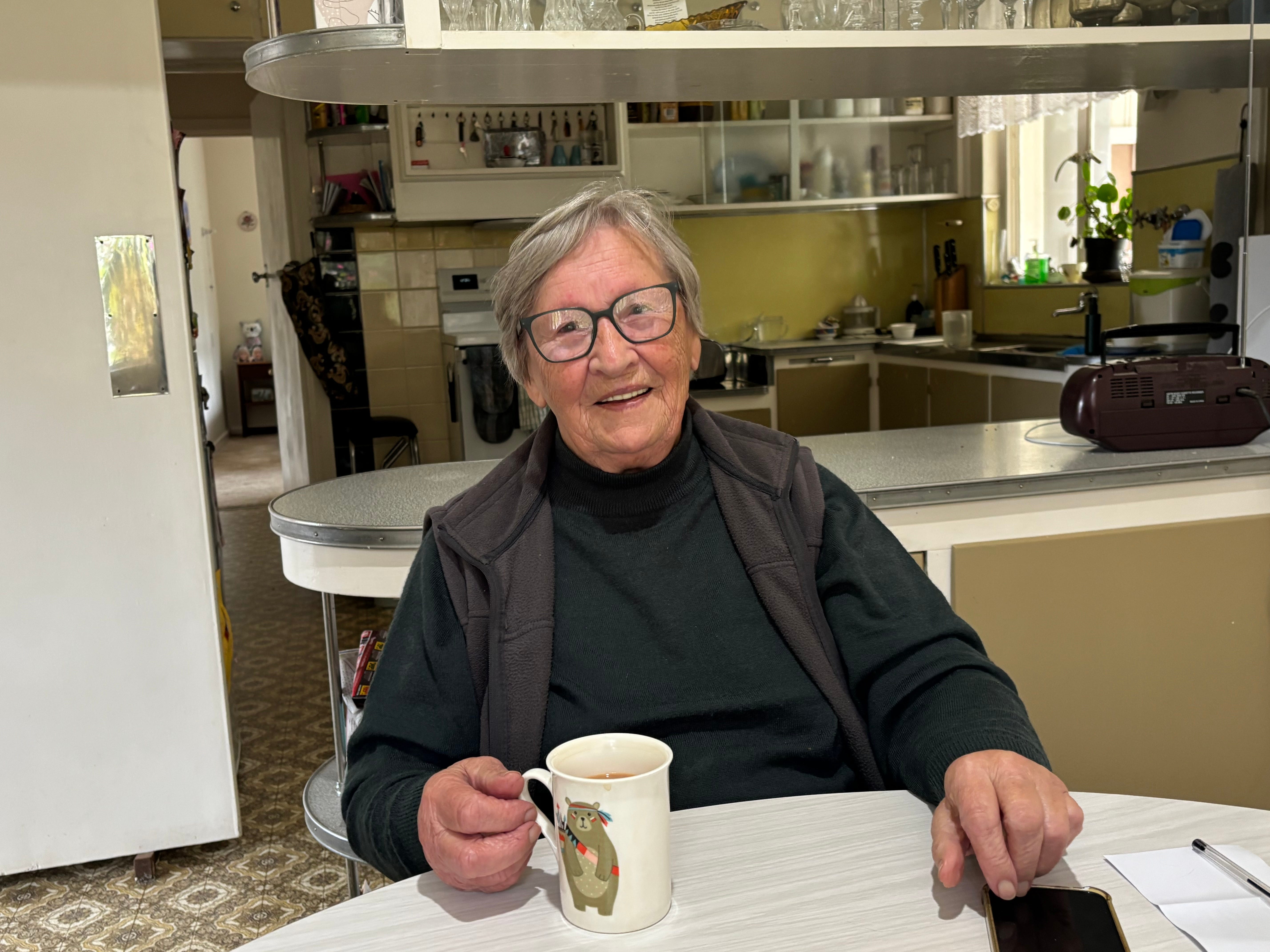 Mira Armstrong sitting at a kitchen table with a mug of tea wearing glasses and smiling at the camera.