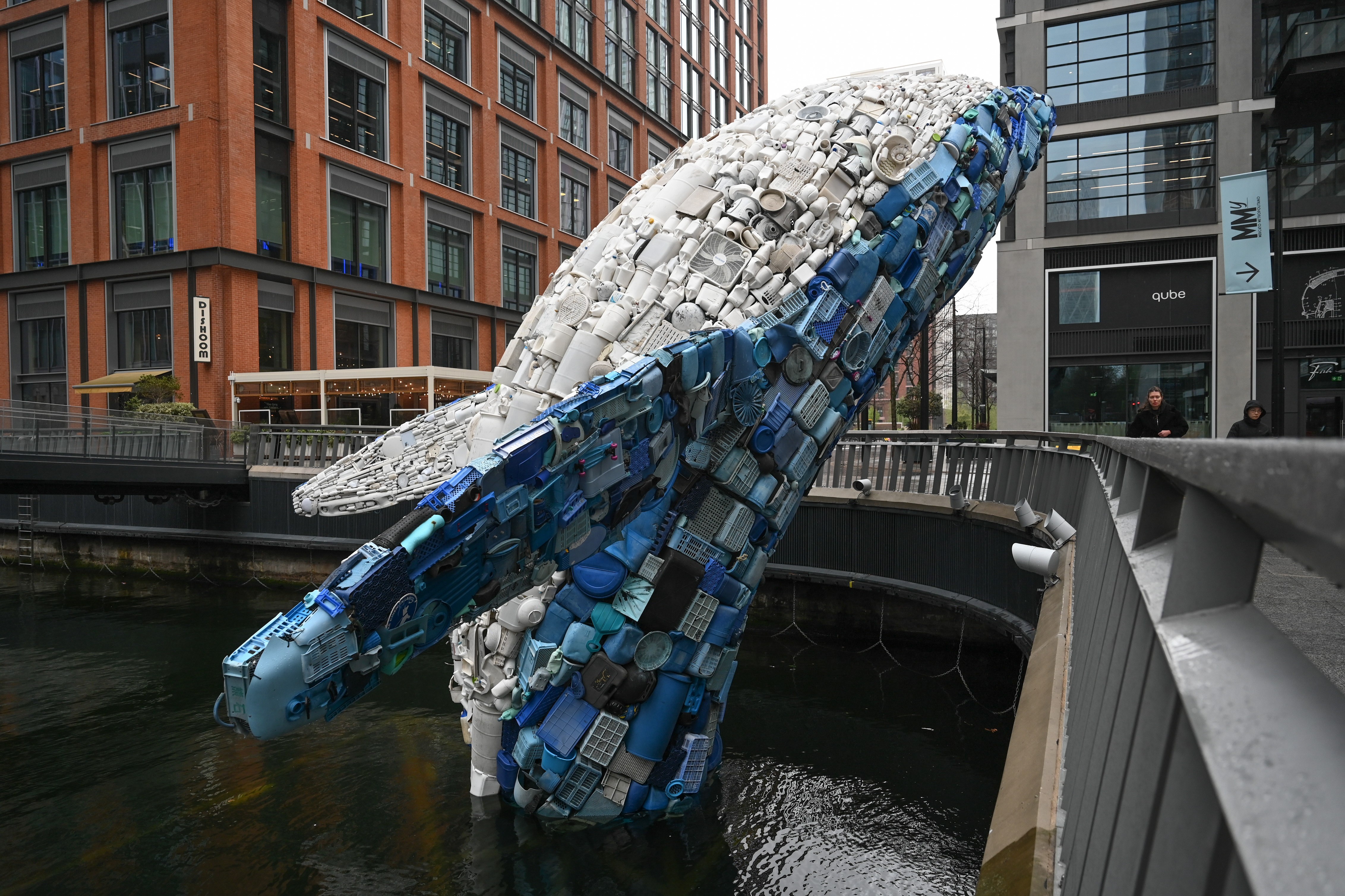 A giant sculpture of a breaching whale, made entirely of pieces of plastic waste, standing in a wharf area, near buildings and walkways