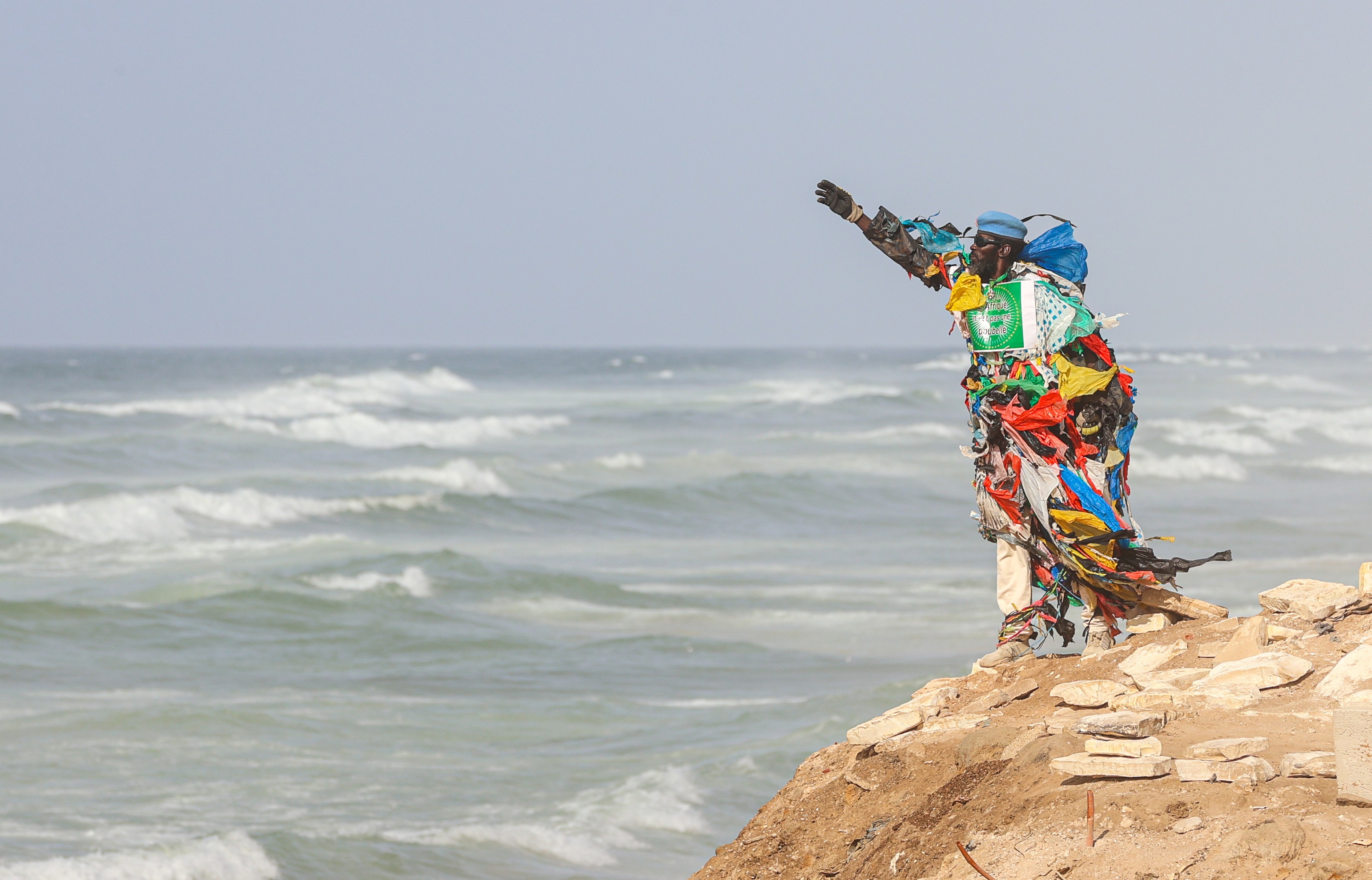 A man wearing a costume made of hundreds of plastic bags stands on a shoreline with his arm in the air.