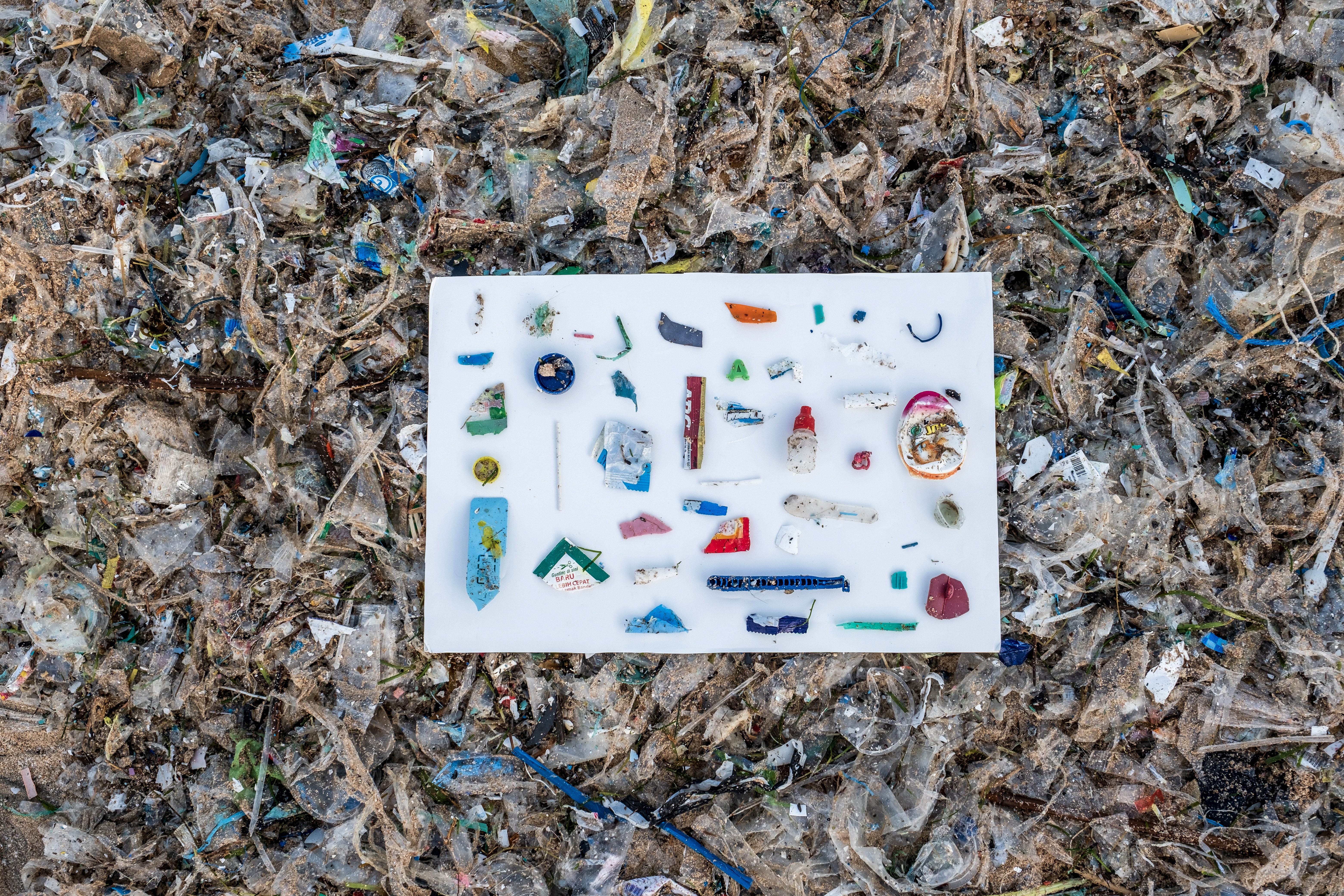 A white board has been placed on top of a pile of plastic waste. On the board are arranged several dozen individual plastic items, including a candy wrapper and part of a disposable razor.