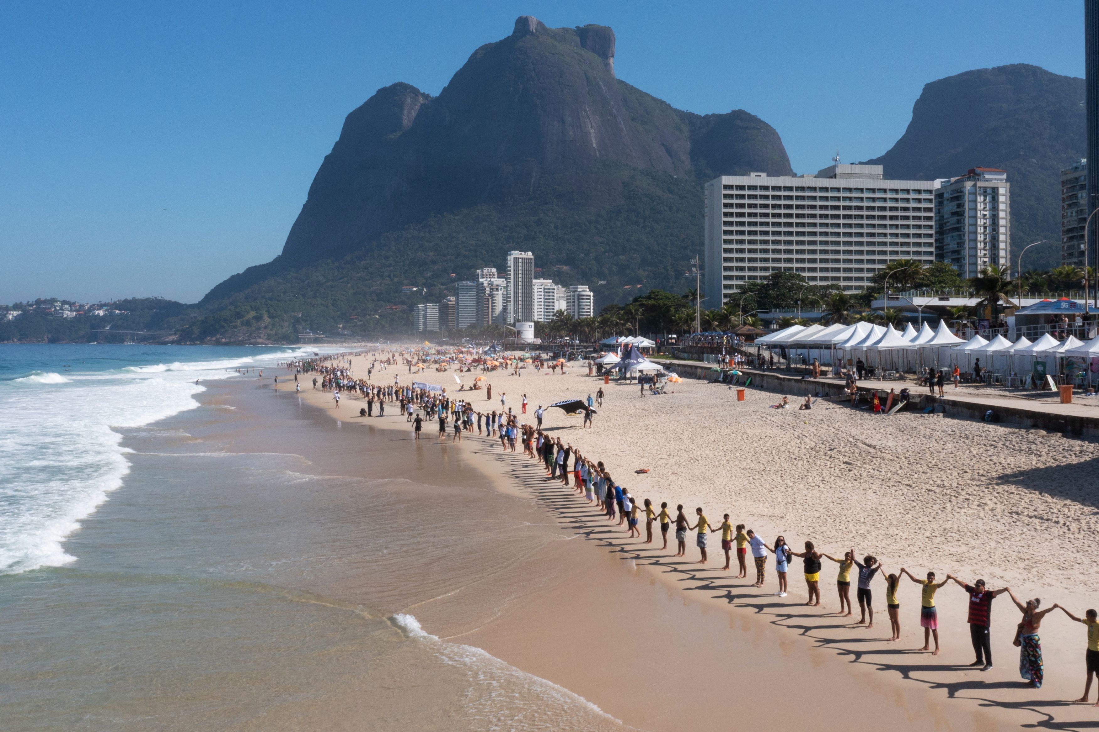 A long line of people stand side-by-side, holding hands on a beach in Rio de Janeiro.