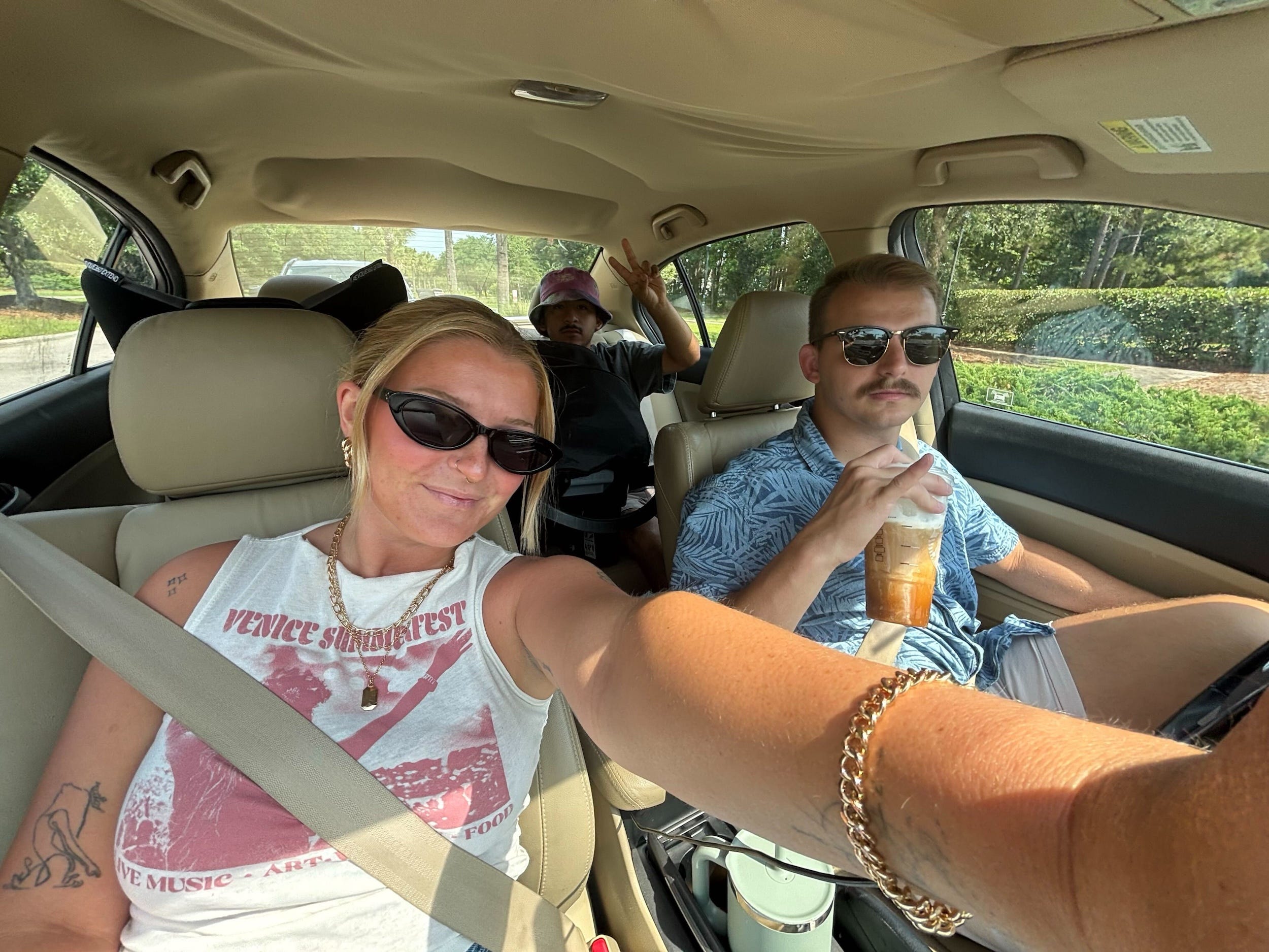 A selfie of a woman and two men in a car. One man sits in the backseat.