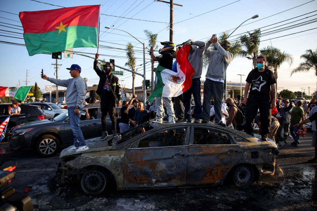 Protestors on a burned-out car, holding flags.