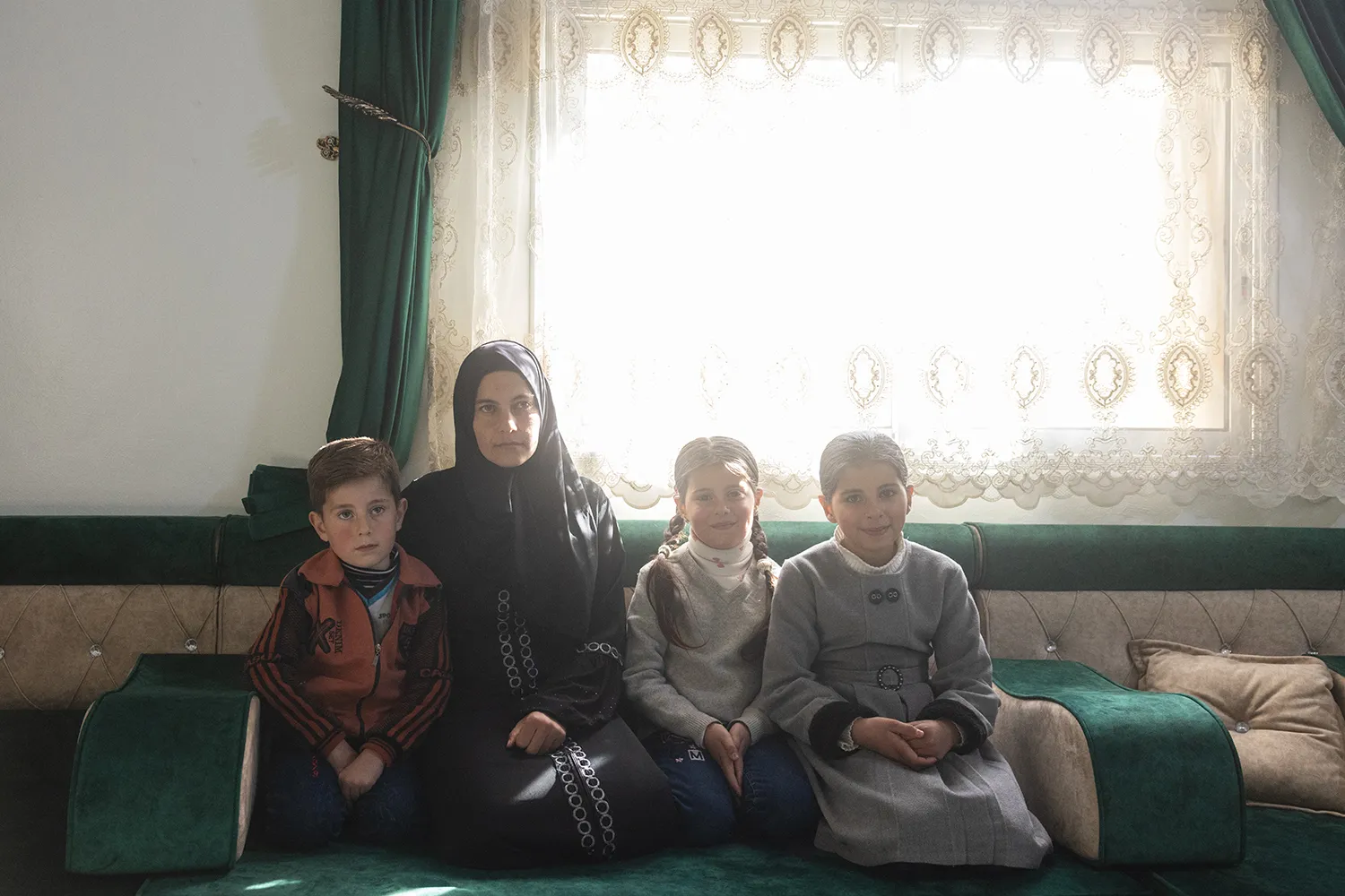 A woman in a black dress and headscarf sits with her three children on a couch in front of a opaque white lace curtain, backlit by the bright light streaming through the fabric. The children appear to range in age from roughly 4 years old to roughly 10 years old.