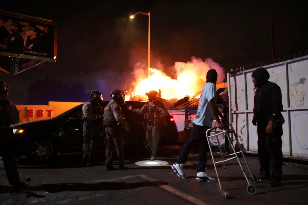 A fire burns in the background as law enforcement officers assist a man during a protest in Los Angeles.