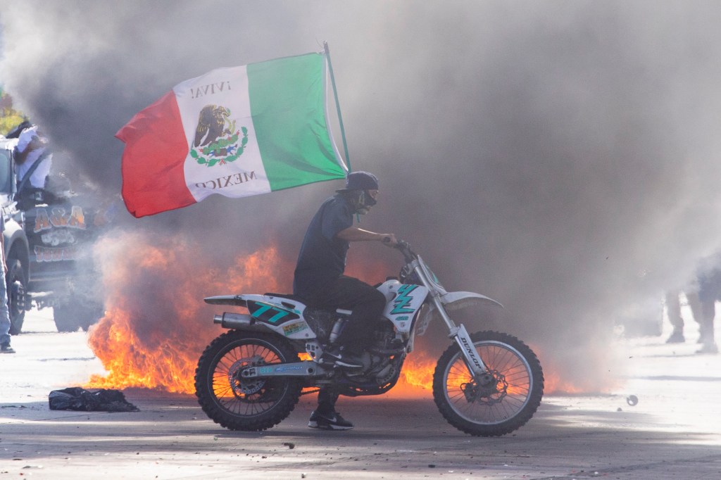 Person on a motorcycle with a Mexican flag during a protest with fire.