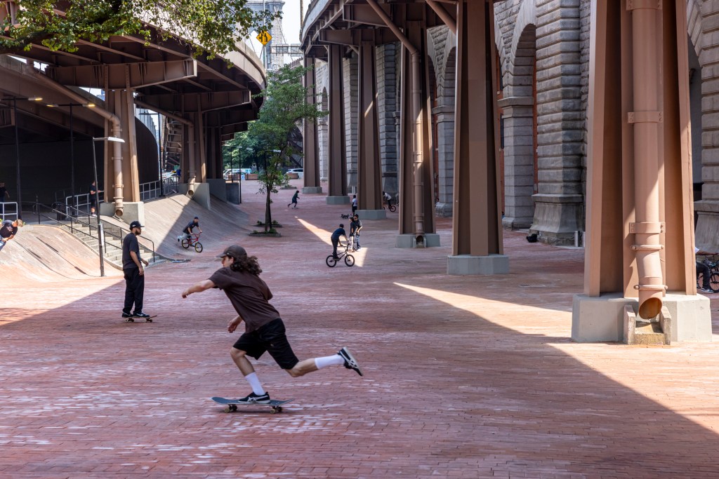 Skateboarders and BMX bikers at a plaza under a bridge.
