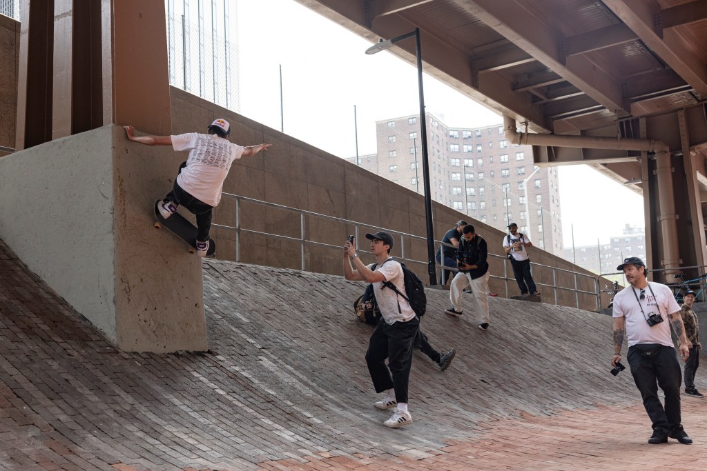 Skateboarder performing a trick under a bridge, with several onlookers taking photos.