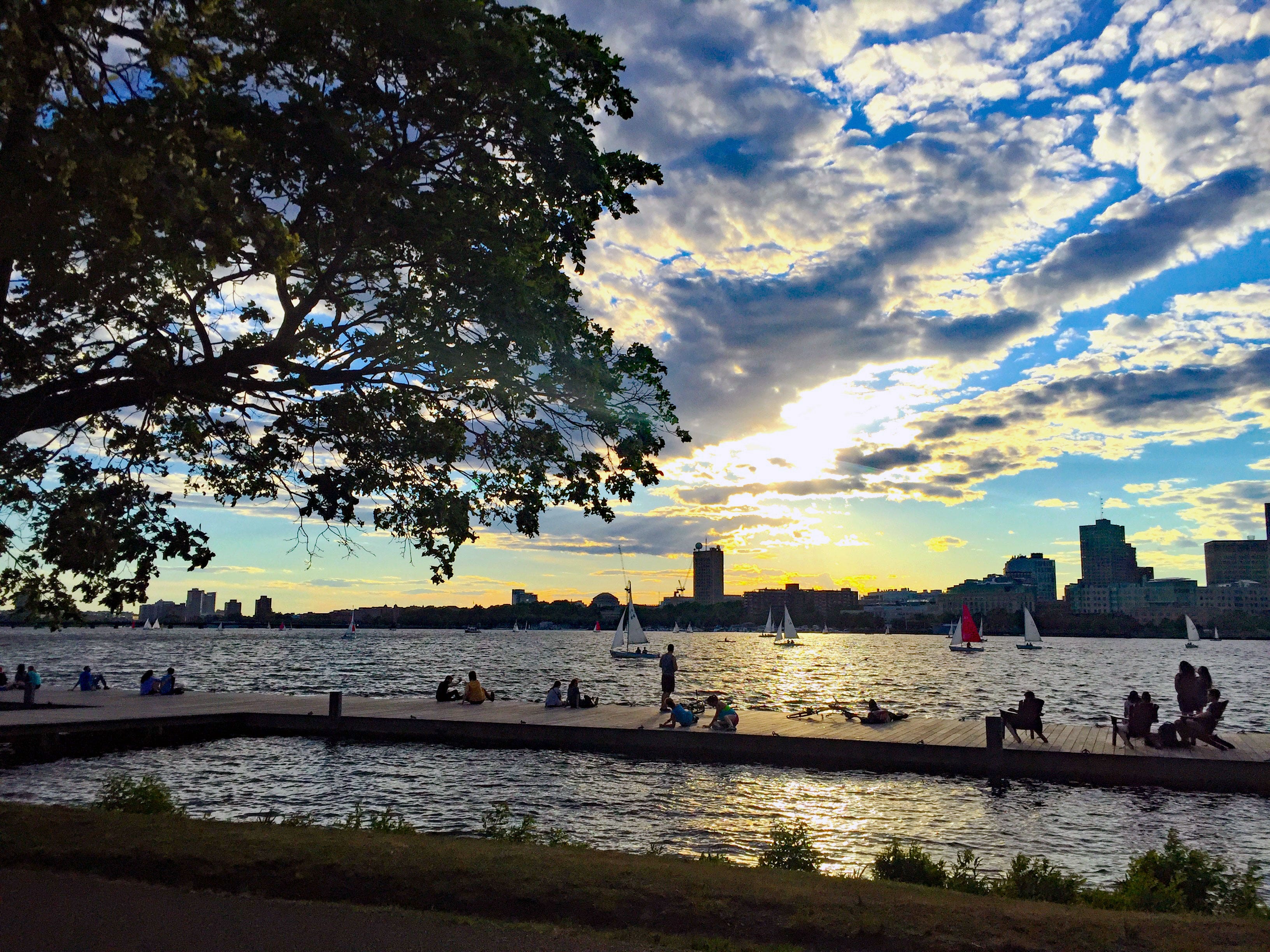 People sitting on a pier looking out at boats and tall city buildings at sunset.