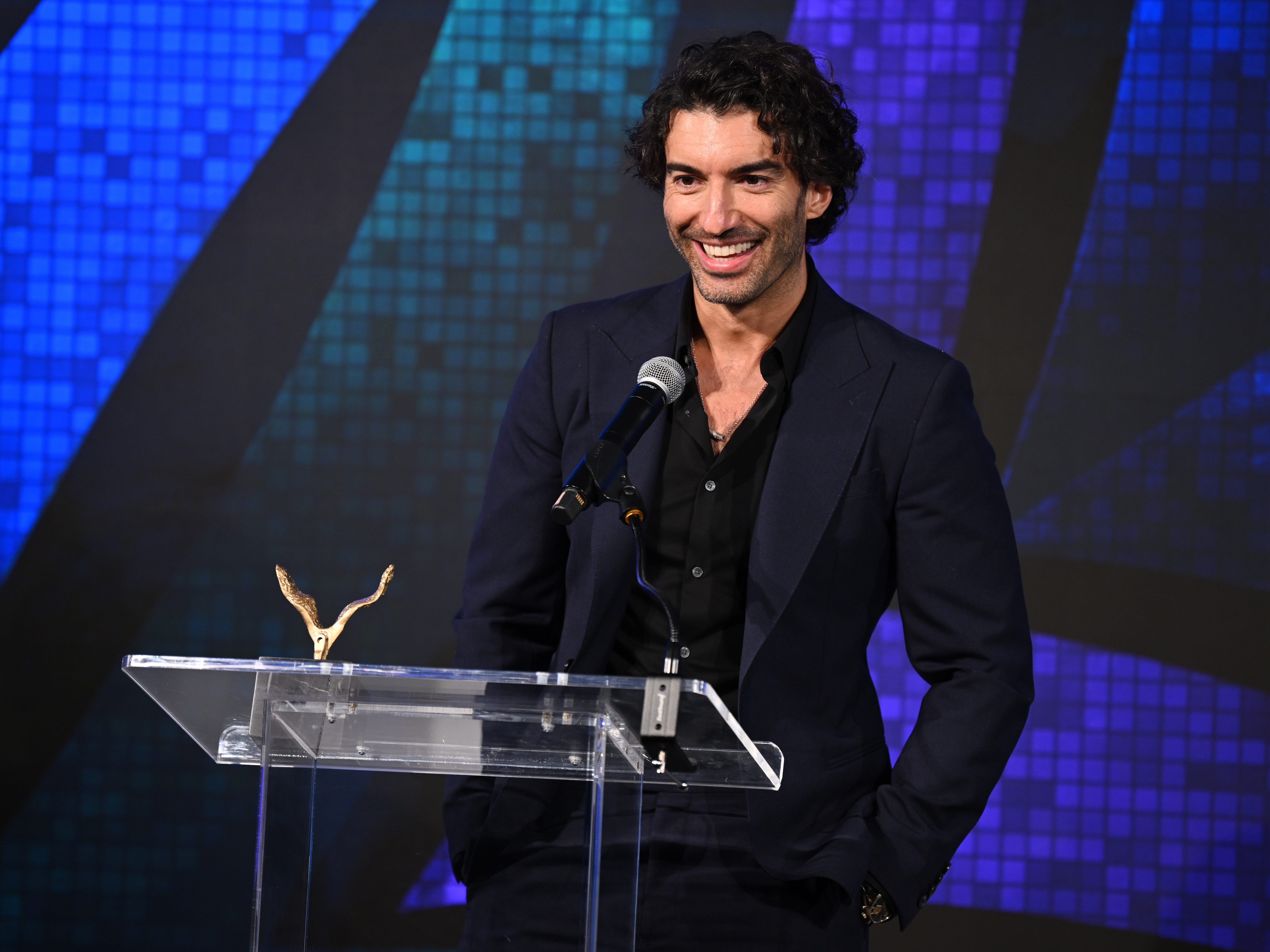 A picture of Justin Baldoni in a suit standing behind a podium with a mic and a gold bird-like award on it.