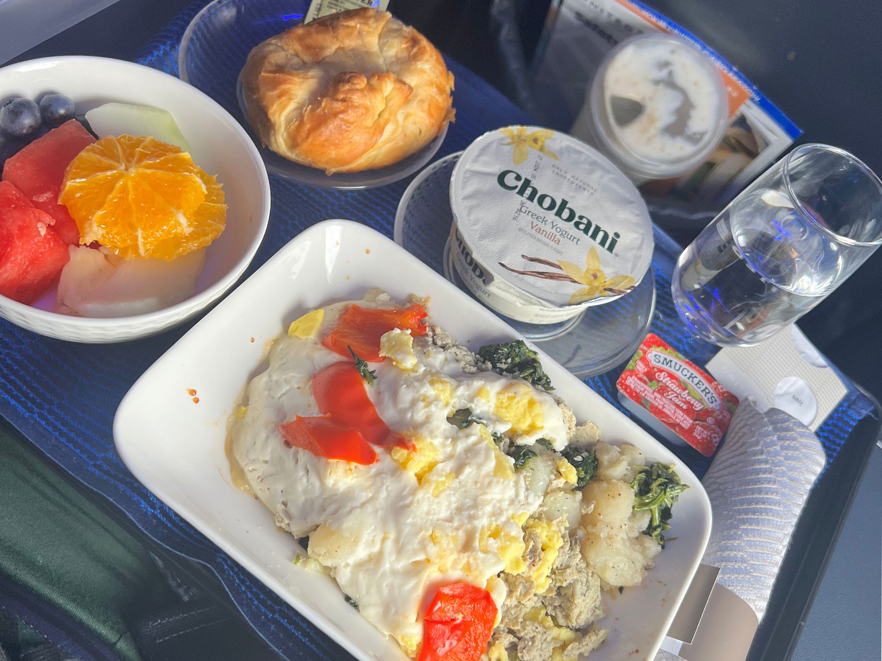 A bowl of fruit, pastry, Greek yogurt, omelette, glass of water, and package of strawberry jam on a tray.