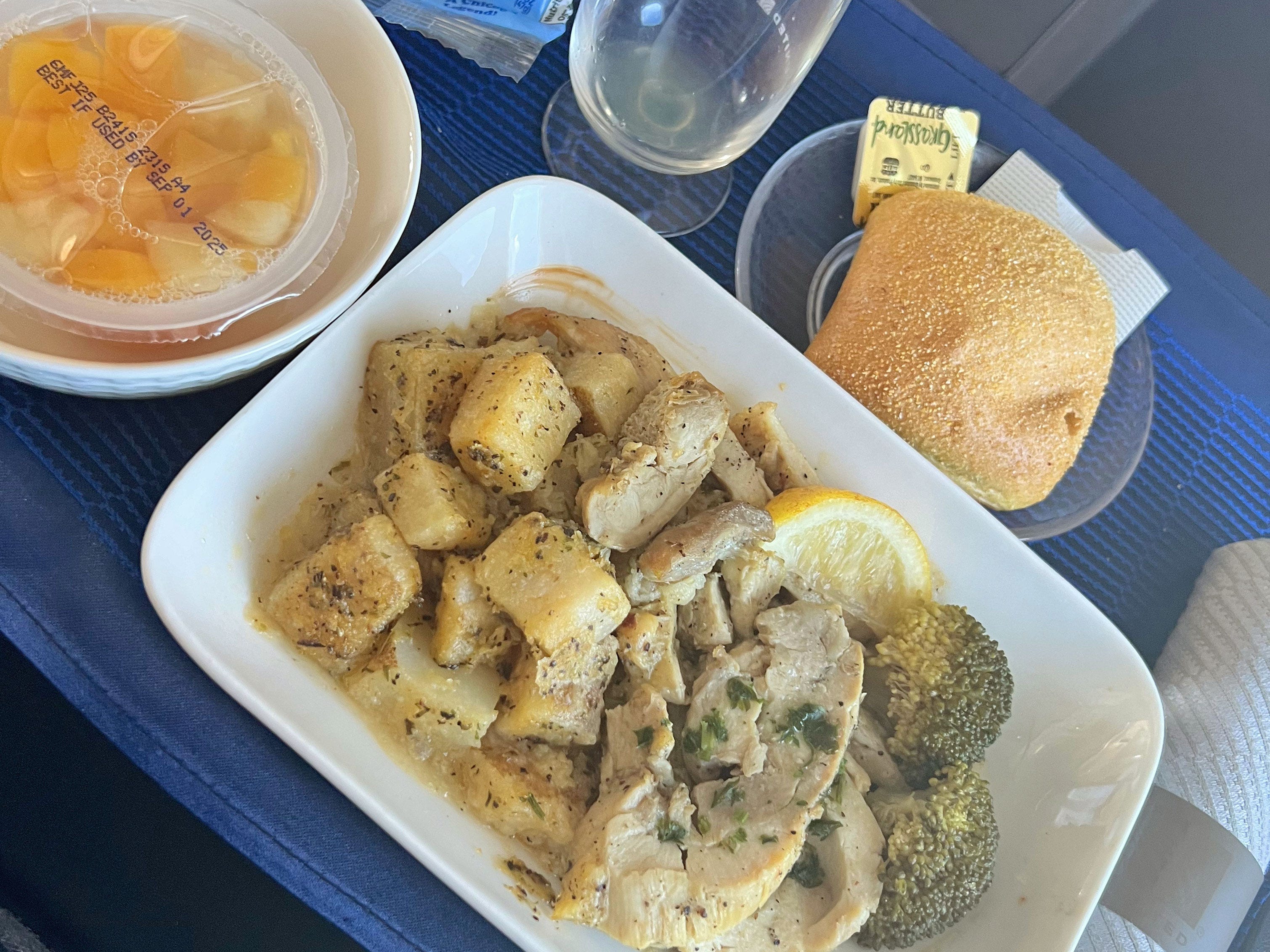 Chicken with potatoes and broccoli, a cup of fruit, and a roll with butter on a tray.
