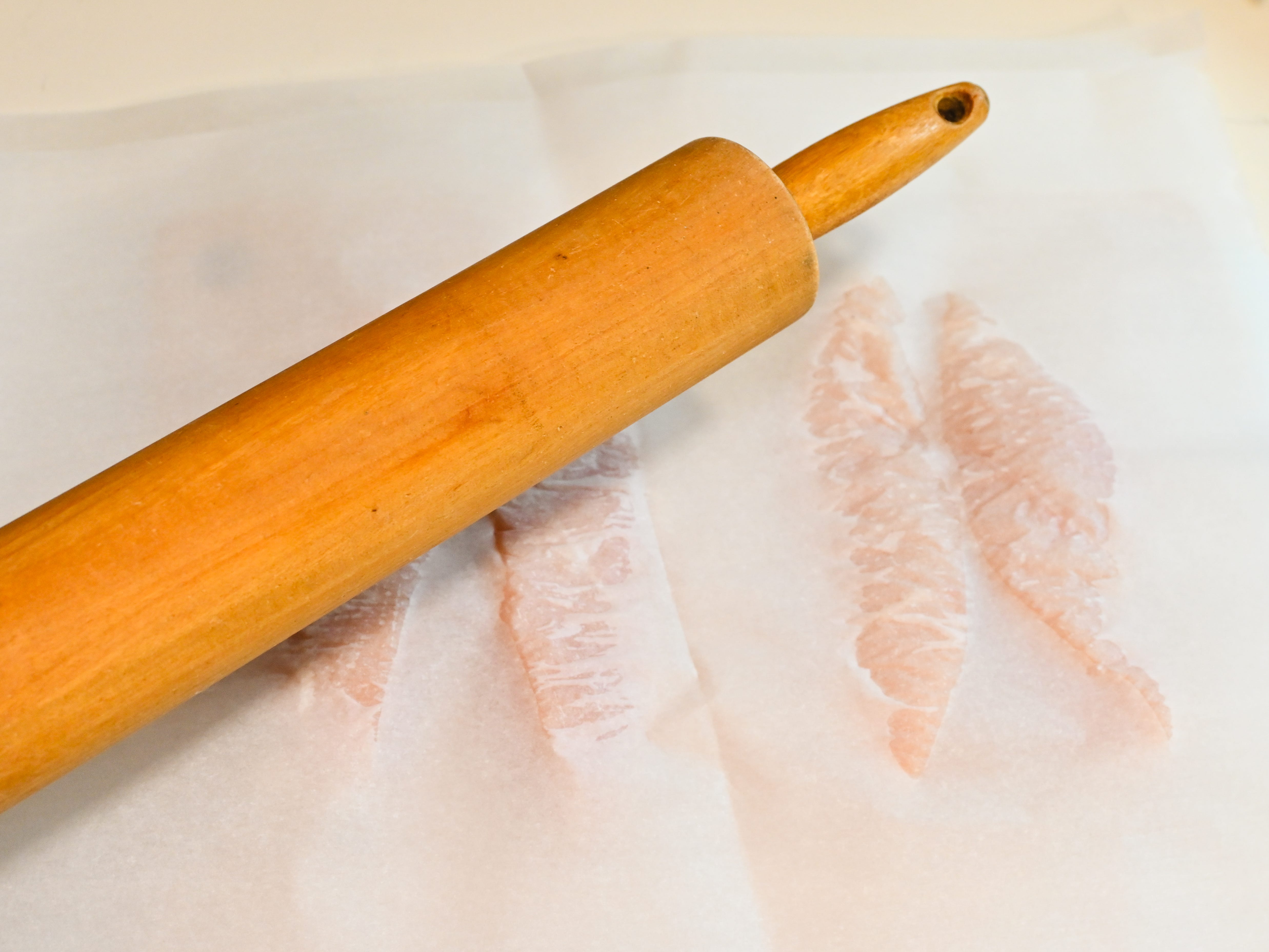 The sliced chicken breasts are sandwiched between two pieces of parchment paper with a rolling pin laid on top.