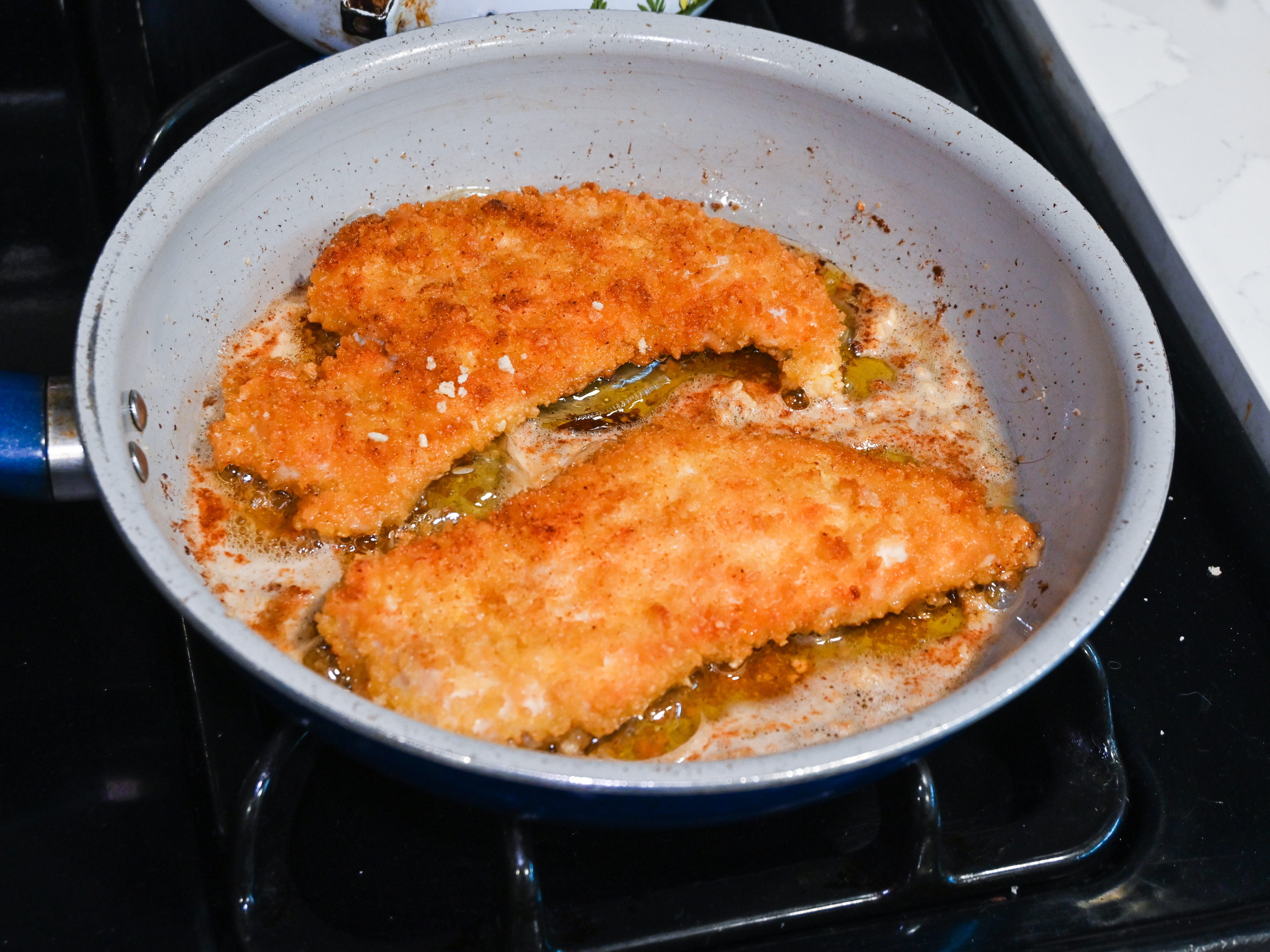 Two chicken breasts cooking in oil on a pan on a stove.