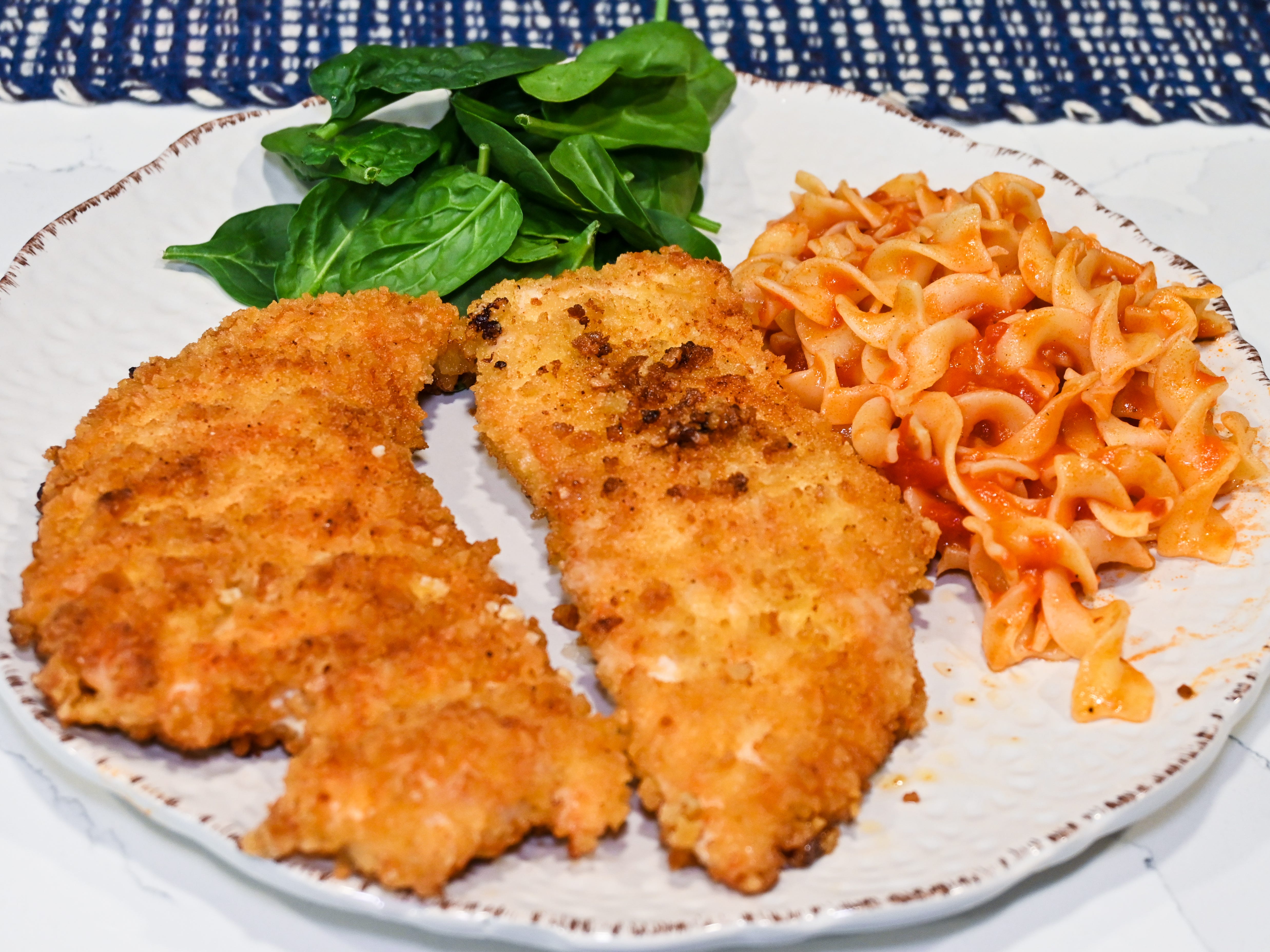 A plate of two cooked chicken cutlets with pasta and spinach leaves.