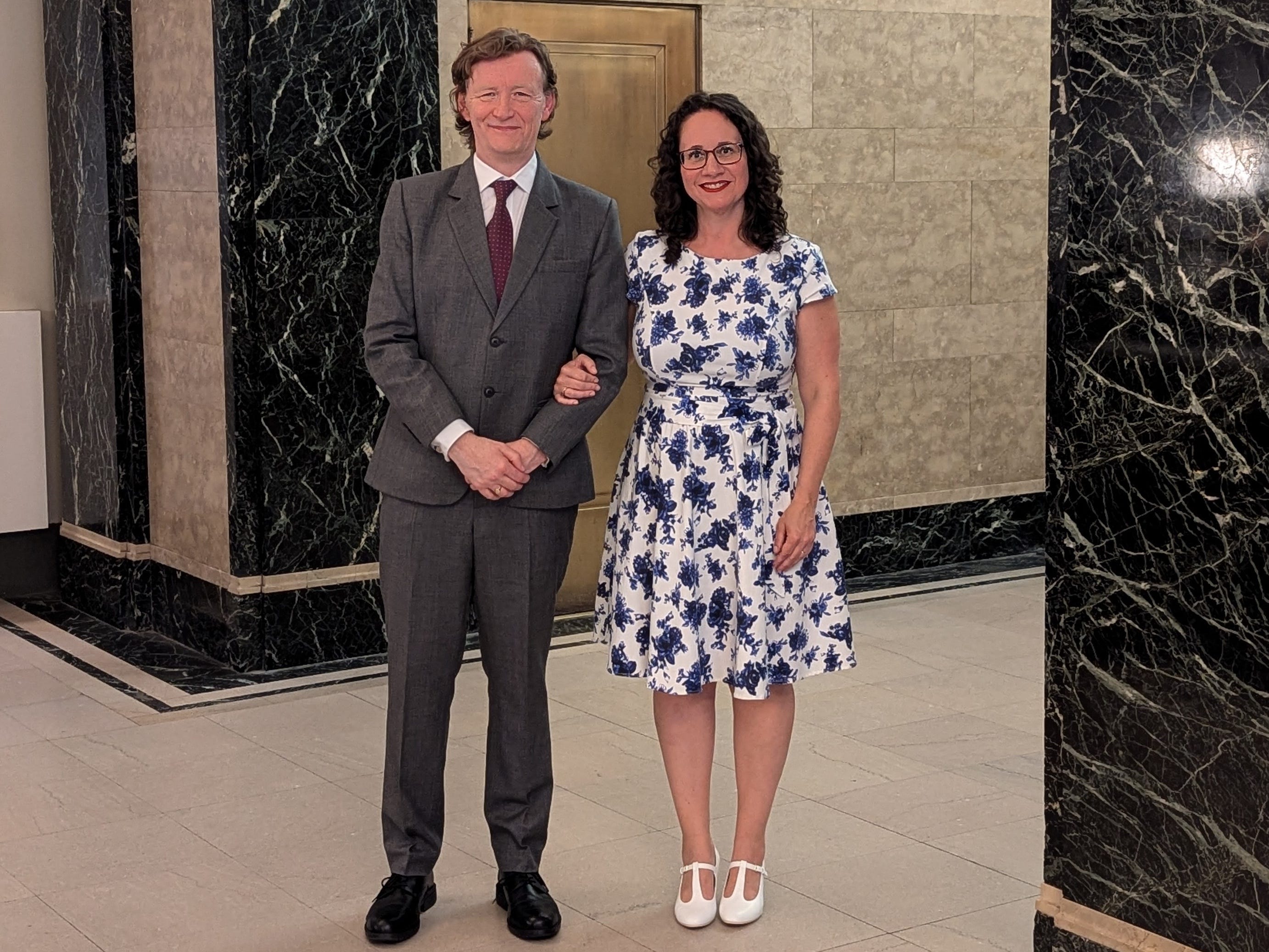 A man in a suit and a woman in a blue and white dress stand arm-in-arm at NYC's City Hall.