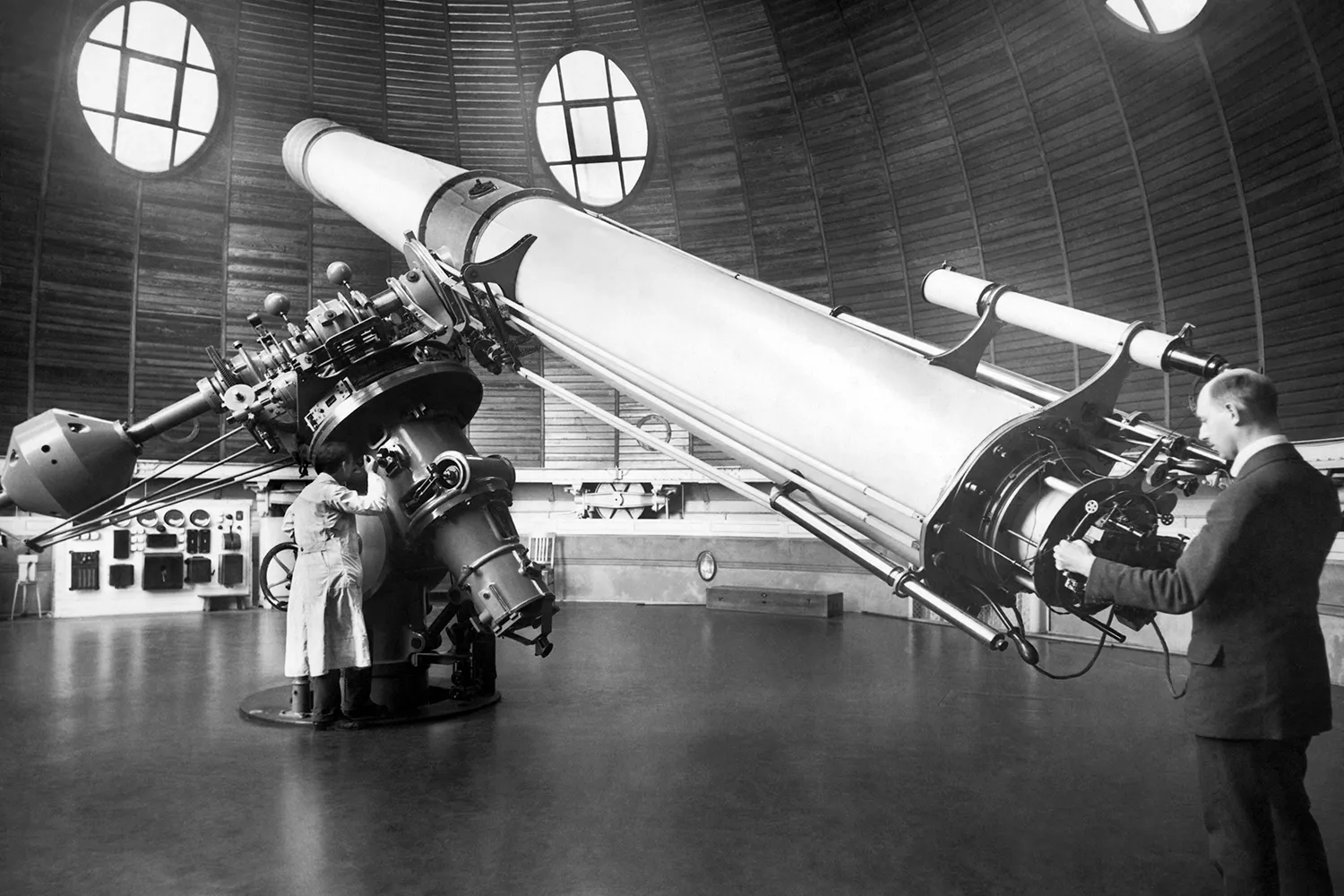 In this black-and-white historic photo, two men operate a massive telescope positioned in a round observatory building. One man looks through the eyepiece while the other operates the base that the telescope rests on. The telescope looks to be about as long as a schoolbus.