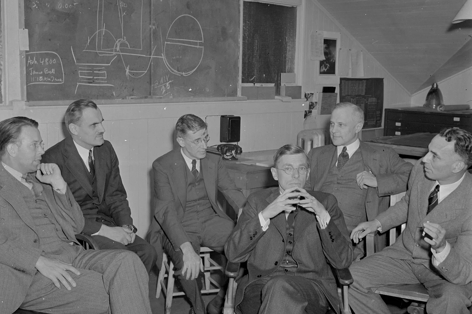 This black-and-white photo shows a group of six men in a classroom in the 1940s. James Bryant Conant sits at center, his hands steepled together. A diagram is drawn on a chalkboard in the background.