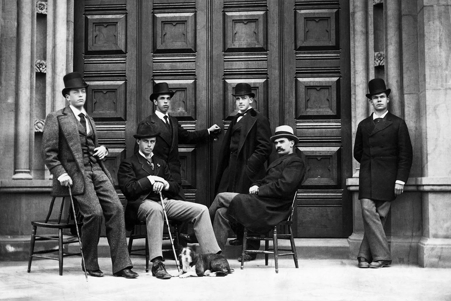 This black-and-white photo shows a posed group of six men in formal coats and hats from the 1870s. Two of the men sit; the others lean against a large wooden door and the columns that surround it.