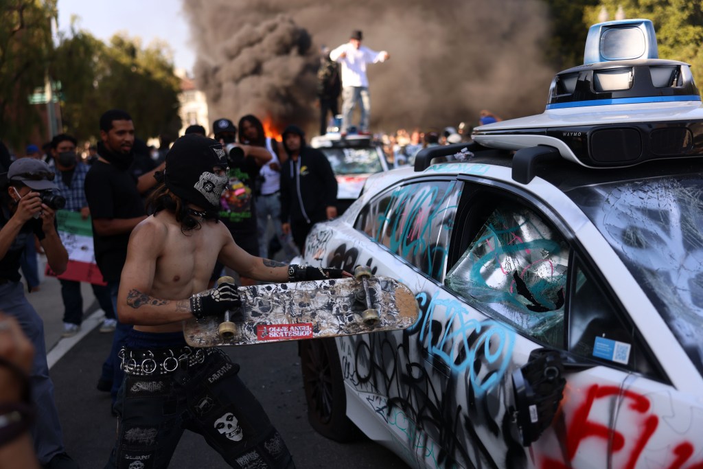 A shirtless man with a mask holds a skateboard during a riot; a vandalized car is in the background.
