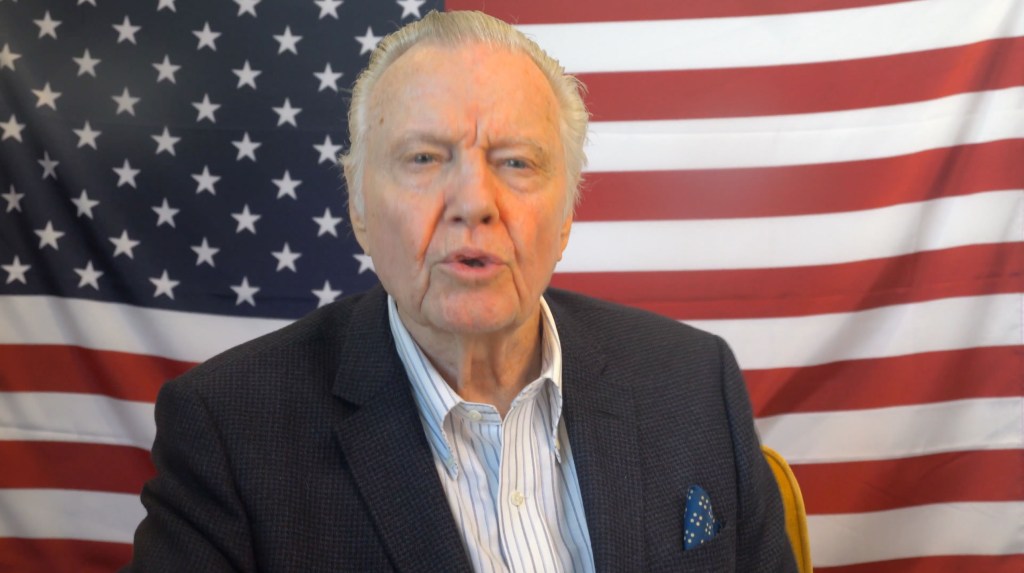 Jon Voight speaking in front of an American flag.