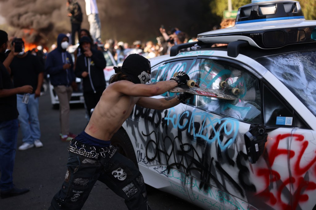 A person smashing a car window with a skateboard during a protest.