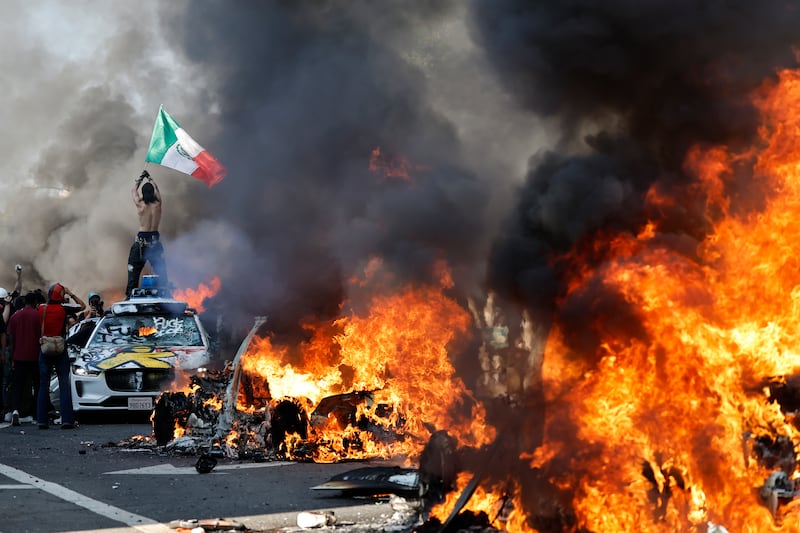 A protestor holds up a Mexican flag as burning cars line the street on June 08, 2025 in Los Angeles, California.