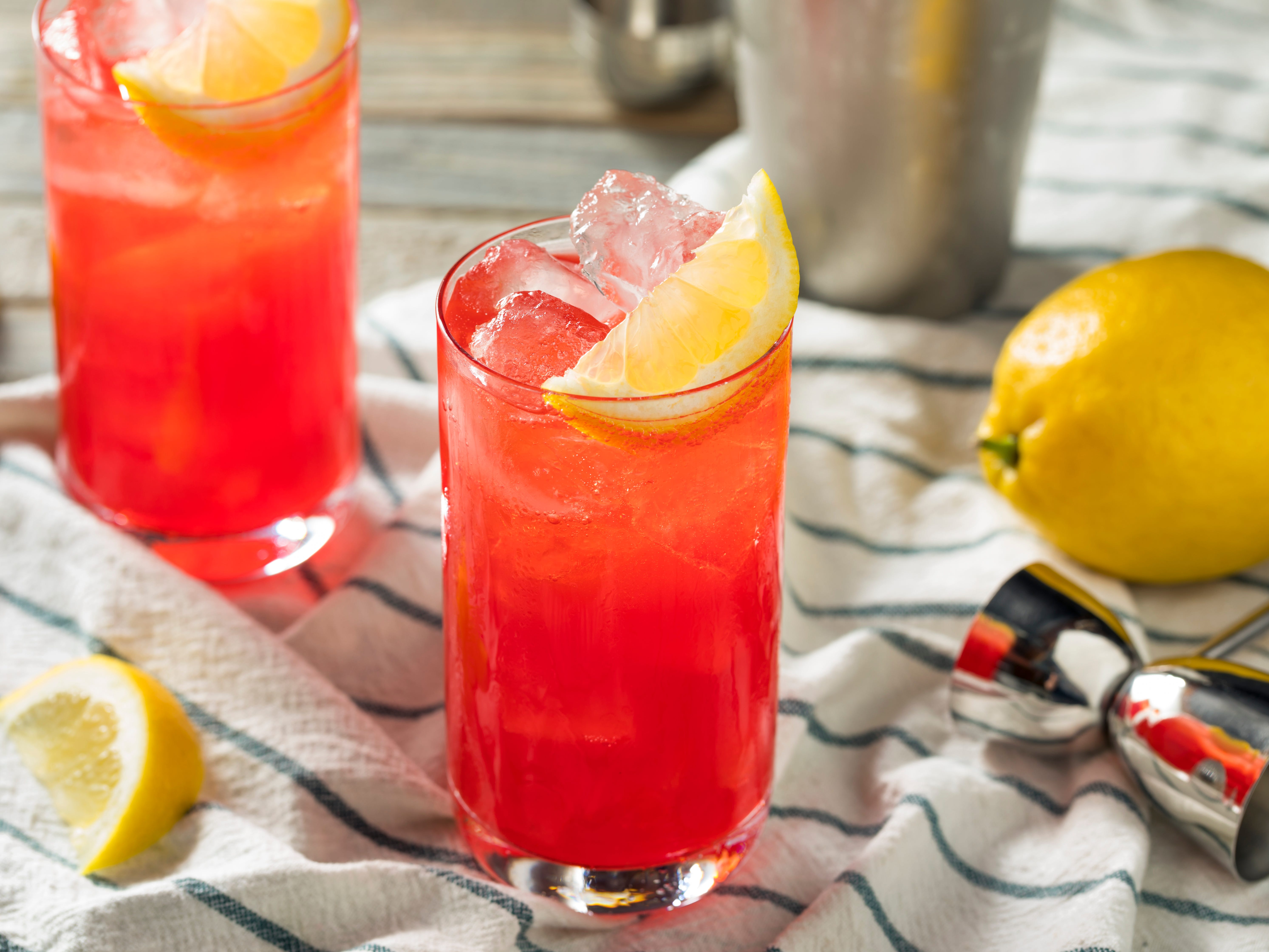 Two glasses of sloe-gin fizz cocktails with lemon slices and ice inside on a white-and-green striped cloth that has bartending tools on top.