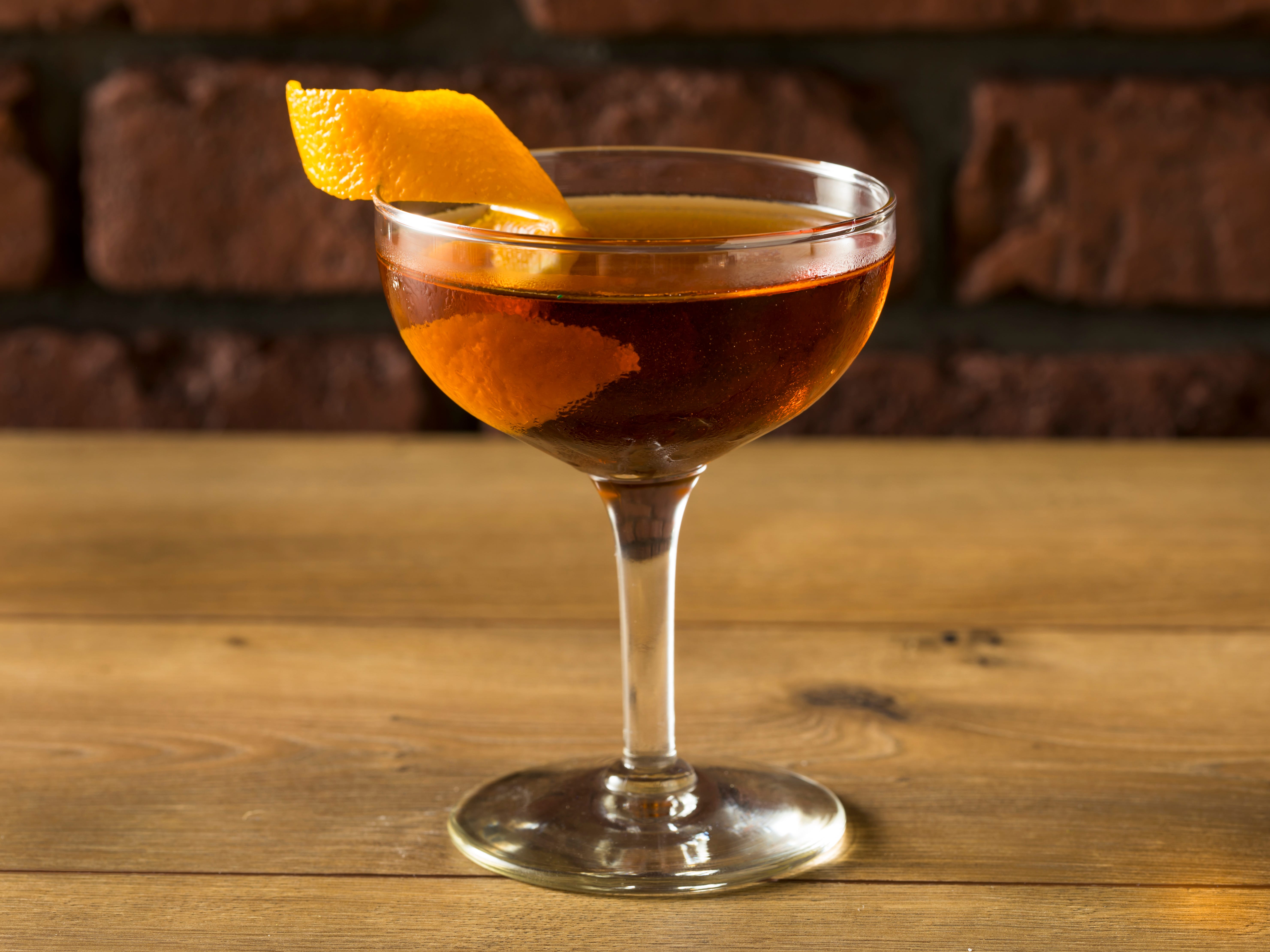A Martinez in a glass with a twisty orange shaving on a wooden surface with bricks in the background.
