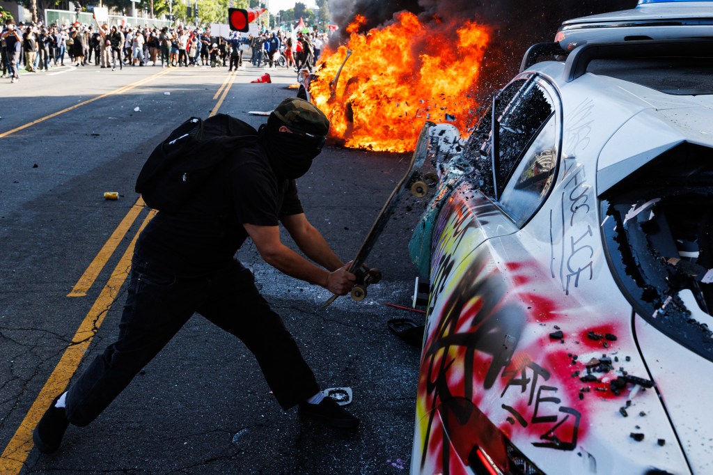 Protestor smashing a burning car with a skateboard during a demonstration.