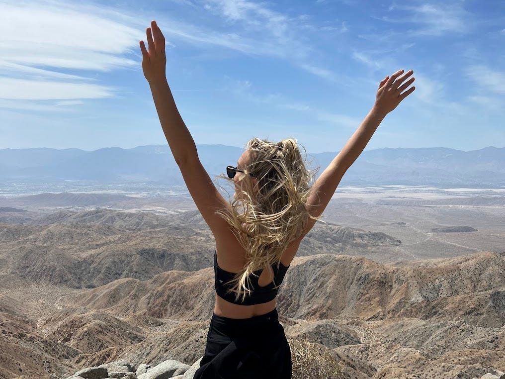 Chloe stands at the summit of a hike in Joshua Tree National Park, raising her hands in the air.
