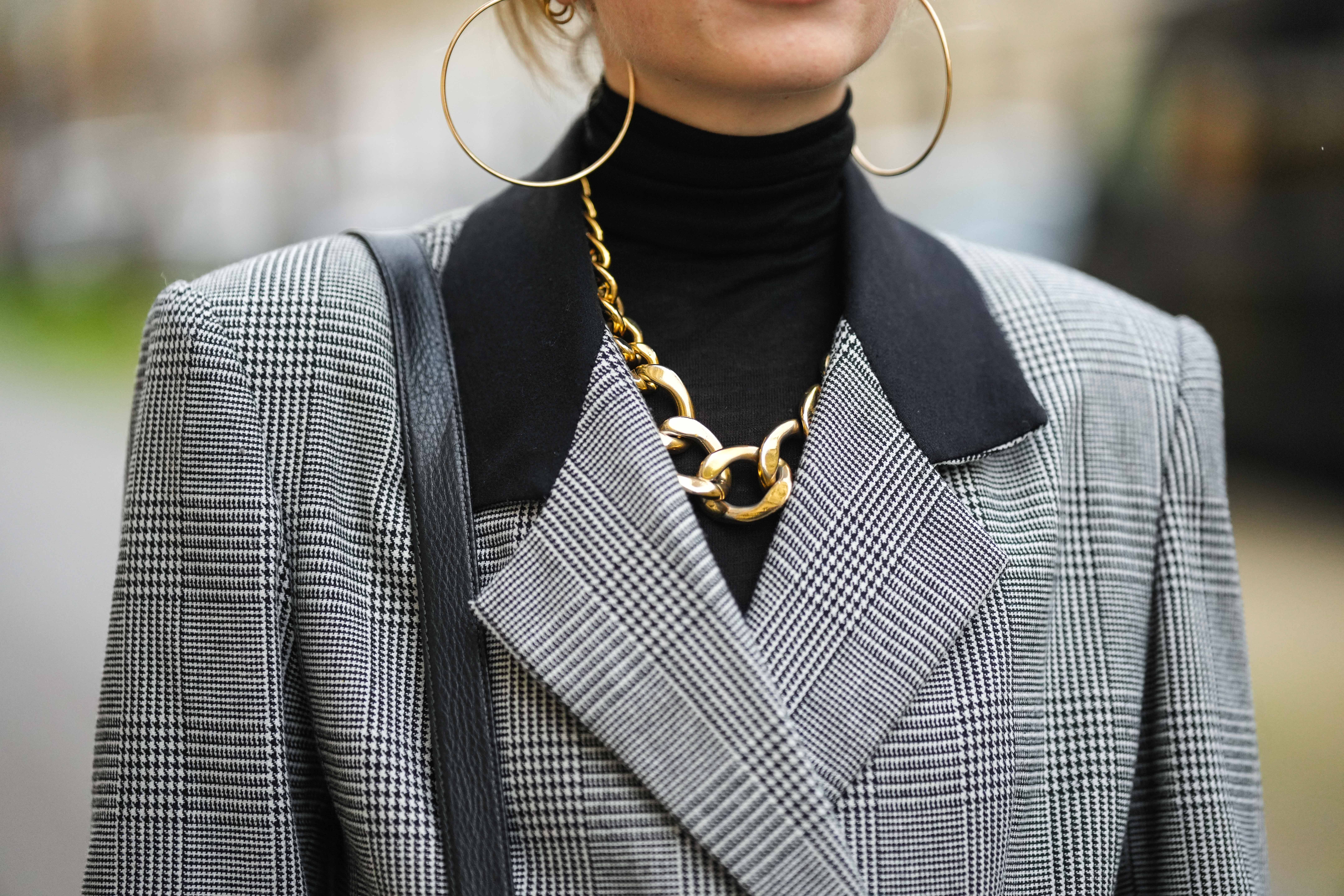 Person wearing gold large earrings, a gold large chain necklace, a black turtleneck with checkered blazer