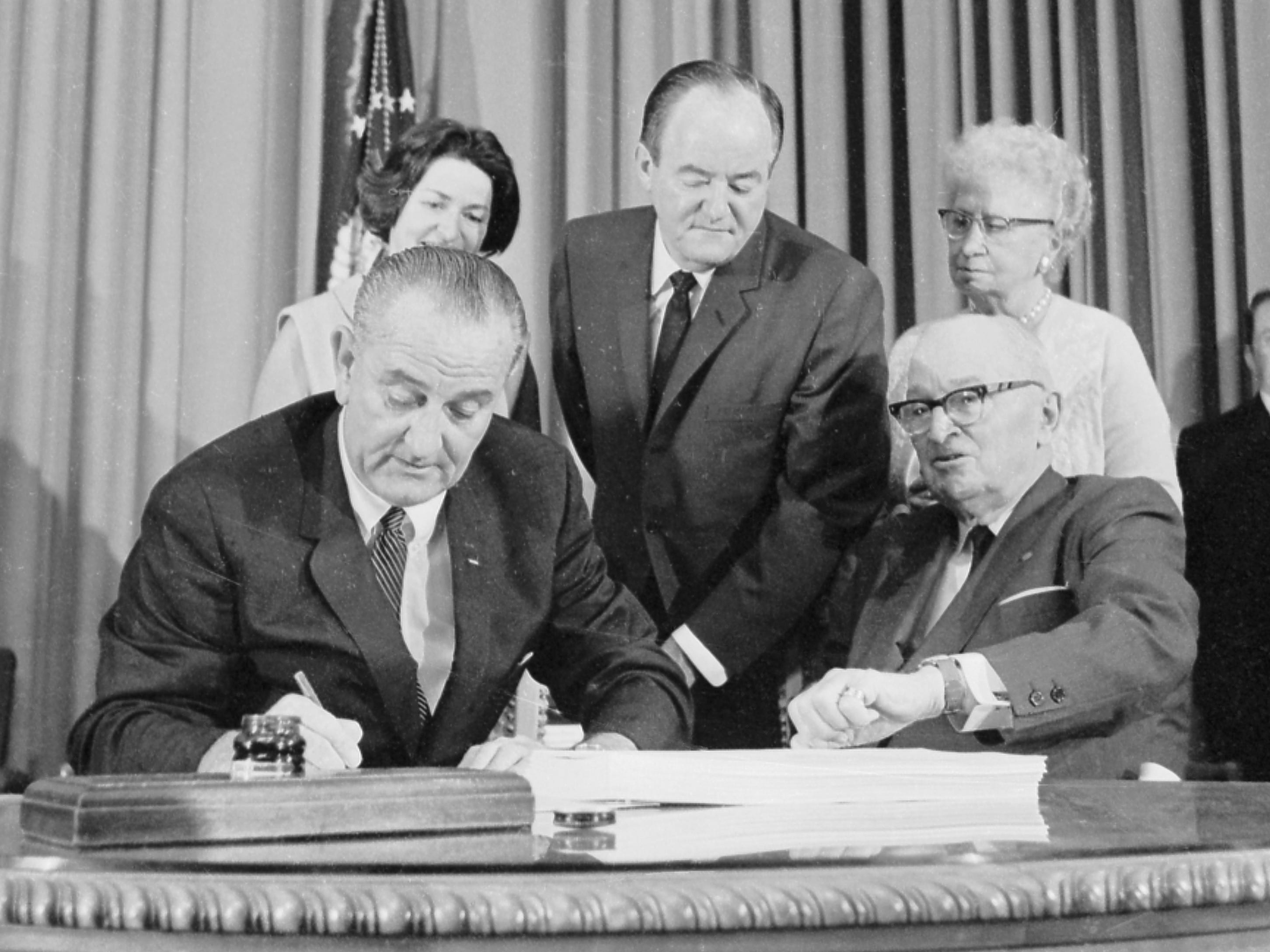 President Lyndon B. Johnson signs the Medical Bill as H.H. humphrey looks at LBJ's watch, and Harry Truman also looks on.
