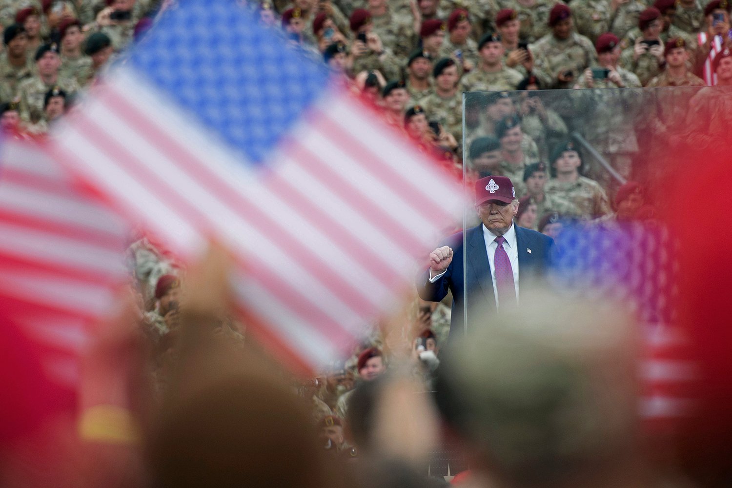 Donald Trump is on stage and lifting his hand in the air. In the background are troops, and an American flag is in the foreground.