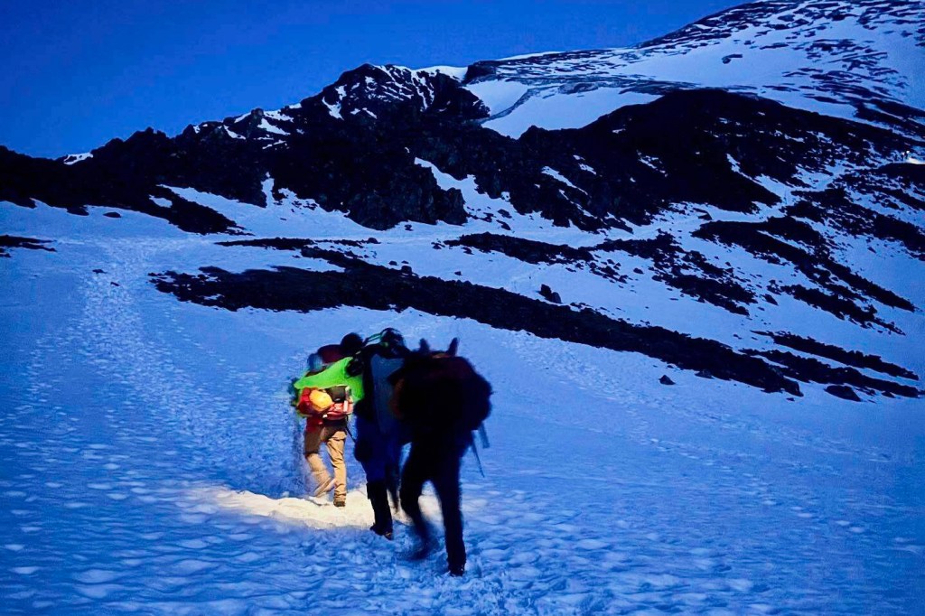 Alpine rescue team members assisting two men down a snowy mountain at night.