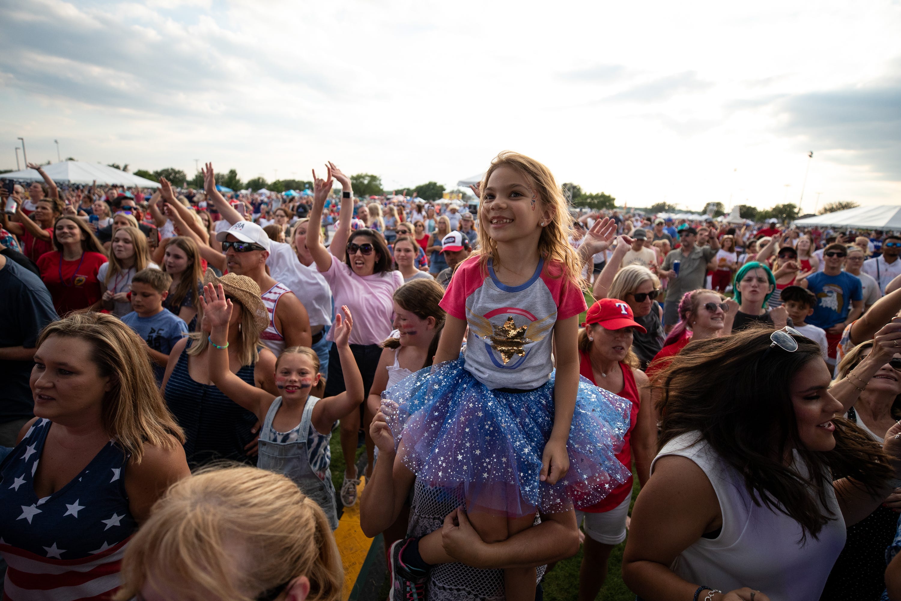 A young girl sits on the shoulders of a man at a community concert. She's surrounded by many other people.