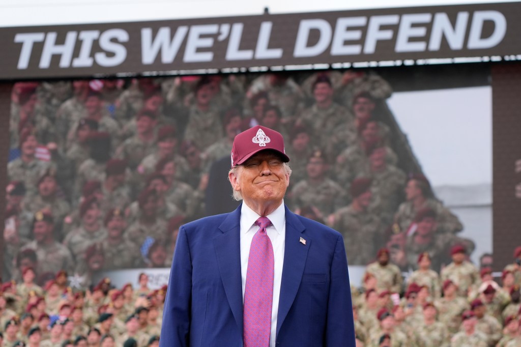 President Trump at Fort Bragg, wearing a maroon hat with military insignia, in front of a screen displaying soldiers and the words 