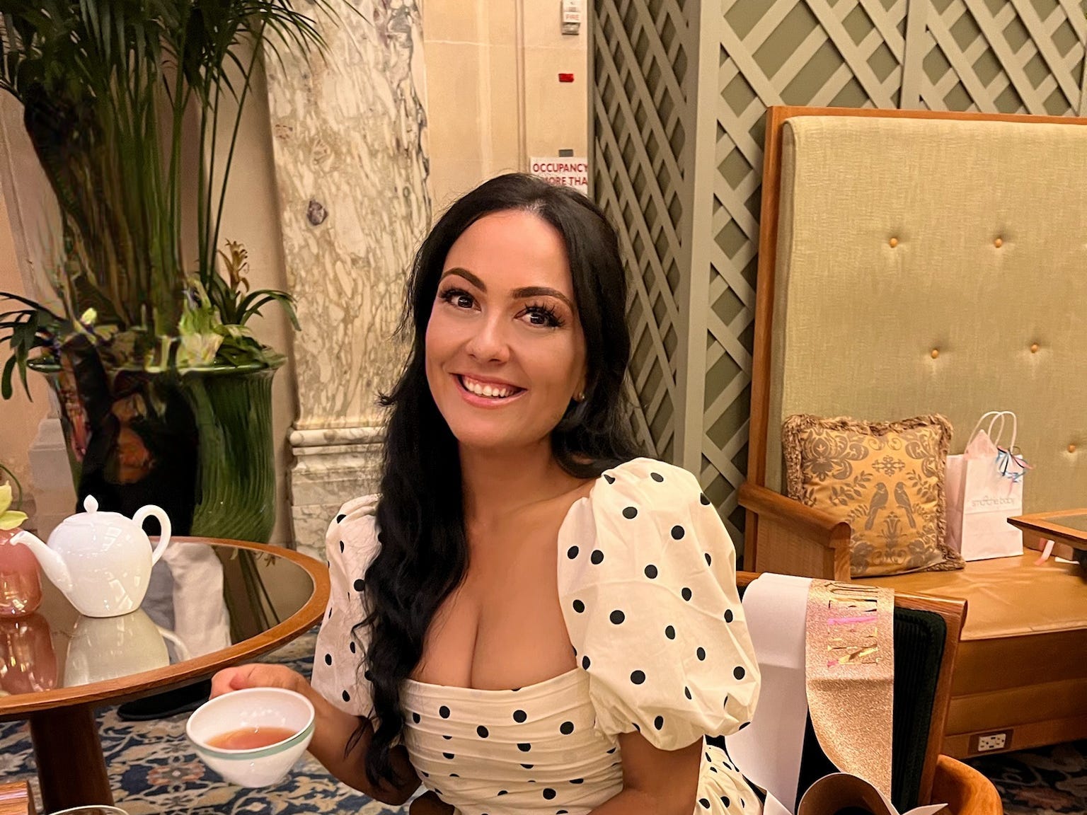 The author wearing a polka dot dress having tea at a brunch table and smiling at the camera.