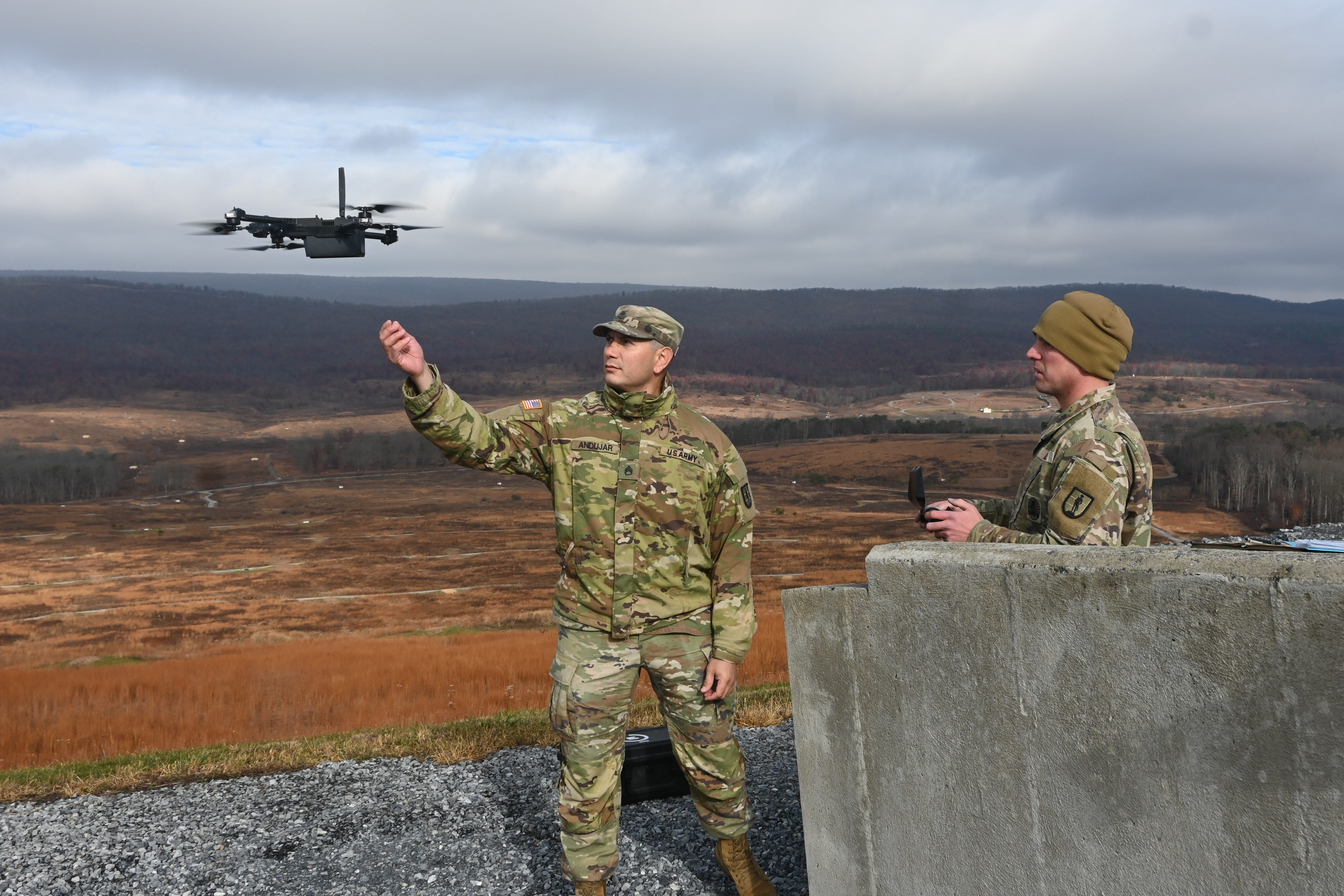 Two men wearing camouflage stand next to a flying quadcopter drone, with rolling hills and a grey sky in the background.