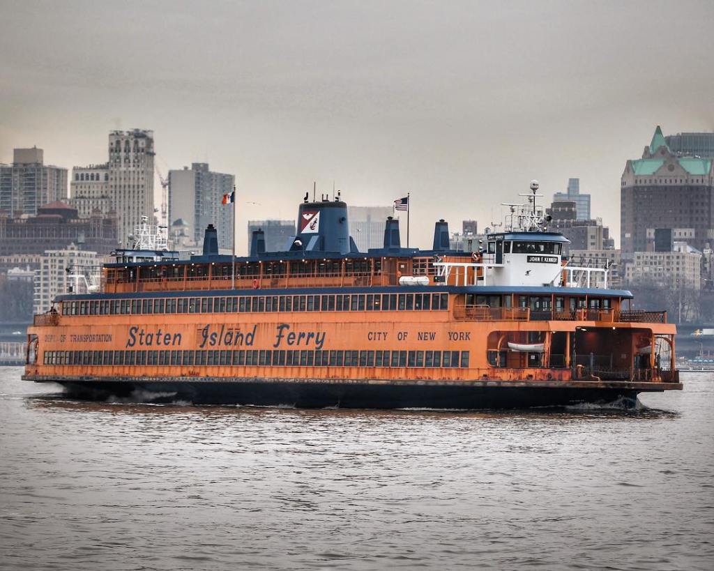 the orange staten island ferry on the water with the skyline behind it