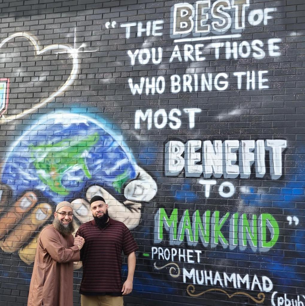 Two men stand in front of a mural that quotes the Prophet Muhammad: “The best of you are those who bring the most benefit to mankind.”