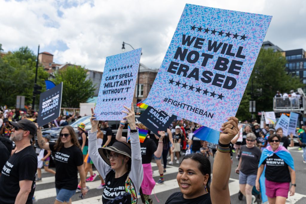 LGBTQ+ veterans march in a Pride parade, holding signs protesting the ban on transgender military members.