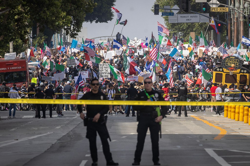 Protesters marching down a street, holding signs and flags, with police officers standing behind yellow caution tape.