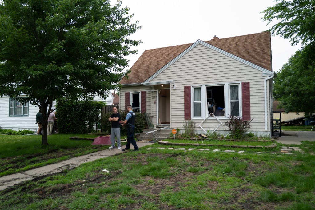 A police officer speaks with a man outside a house with a broken window.