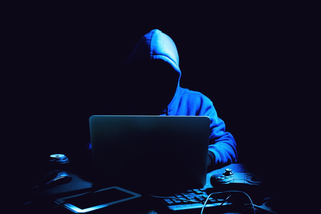 Hooded figure using a laptop in a dark room.
